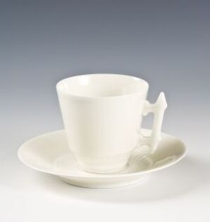  A white feldspathic porcelain tea cup and saucer designed by Porsgrunds Porselænsfabrik AS, featuring a unique element shaped like a miniature chair attached to the saucer, set against a light grey gradient background.