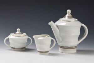  A set of Porsgrunds Porselænsfabrik AS white feldspathic porcelain tableware including a coffee pot, creamer, and sugar bowl, decorated with a glossy overglaze and accented with silver or gray bands near the base, displayed against a light gradient background.