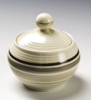  A cream-colored, round feldspar porcelain container with a lid, featuring horizontal brown bands, designed by Porsgrunds Porselænsfabrik AS. The object is set against a light background, with a gentle reflection hinting at its glossy surface.