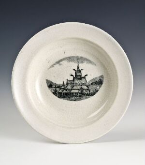  A cream or off-white colored ceramic plate featuring a detailed black and white image of a historic sailing ship at its center, set against a neutral grey background.