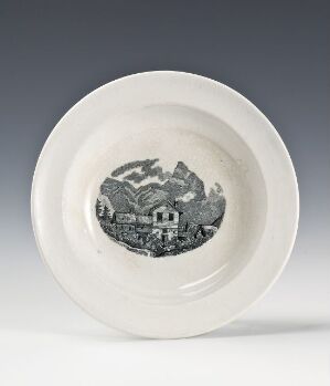  A white circular plate with a monochromatic illustration of a rural village scene at the center, featuring houses among trees and hills. The plate has a simple design with an unadorned rim and a grayscale central image conveying an antique countryside aesthetic.