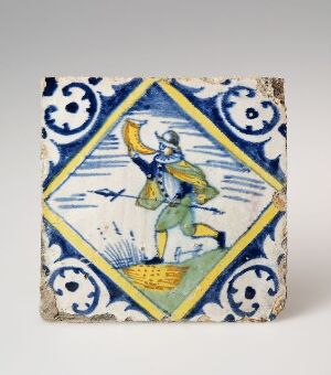  An antique ceramic tile with a hand-painted depiction of a man in traditional attire, possibly a farmer sowing seeds. The tile is edged with a decorative blue and white geometrical and floral border.