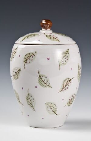  An elegant white porcelain jar with stylized green leaves and small red dots scattered on its surface. The jar has a round, brown knob on the lid, creating a harmonious color palette of white, green, brown, and red.