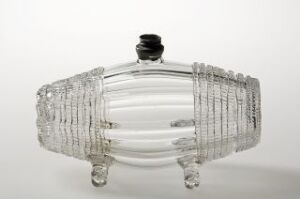  A clear glass sculpture with a ribbed design reminiscent of an inflated tire or lifebuoy, featuring a series of parallel ridges encircling it, three glass feet, and a contrasting black cap on top, set against a plain white background. Artist and title unknown.