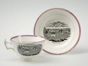  An antique white porcelain tea cup with burgundy trim and a matching saucer, both featuring a grayscale pastoral landscape scene with a large tree and small figures surrounded by cottages and hills.