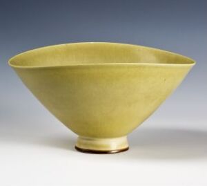  An elegant, wide conical-shaped bowl with a smooth, muted yellow surface standing on a short, narrow base against a neutral grey background. Artist name and title are unknown.