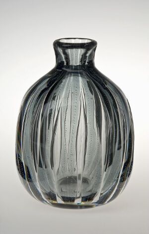  A clear glass vase with vertical grooves creating a textured surface, set against a light grey background. The vase has a rounded body with a tapered neck and fluted opening.