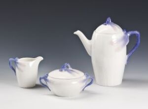  A Rörstrands Porslinsfabrik AB tableware set titled "Trollslända," consisting of a white feldspar porcelain teapot, sugar bowl, and creamer with blue accents on knobs and handles, set against a soft grey background.