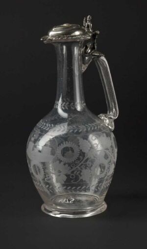  An antique transparent glass pitcher with intricate floral etchings, equipped with a polished silver metal lid and handle, set against a dark background.