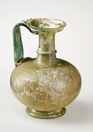  An antique glass jug with a bulbous body and single handle, showing a muted gold color with patches of green and cloudy white, suggesting age and historical significance.