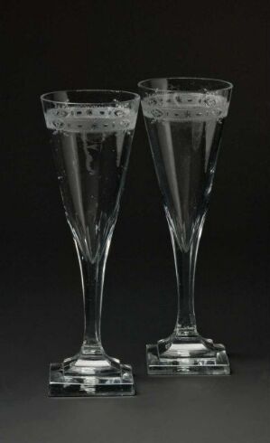  A pair of clear glass goblets with elegant stems and square bases set against a dark background, featuring delicate etching near the rims. Artistname and title are unknown.