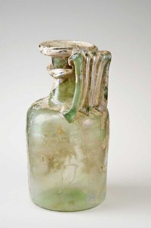  An ancient-style glass vessel with a mottled pale green color and a thick, irregular handle attached to its neck, displayed against a neutral background. The body of the vessel shows subtle variations in color, appearing weathered or aged, while the irregular rim and handle suggest long-term exposure to natural elements.