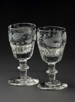  Two intricately etched glass goblets with detailed patterns, standing side by side against a dark background, reflecting light and casting subtle shadows.