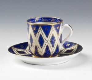  A delicate porcelain cup and saucer set with a cobalt blue base color, adorned with hand-painted white geometric patterns and gold accents, crafted by Porsgrunds Porselænsfabrik AS. The design suggests a luxurious, formal style of tableware.