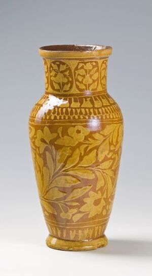  A mustard yellow vase with darker yellow Art Nouveau-style floral patterns, featuring a slender tapered shape with a broad body and a flared lip against a light gray background.