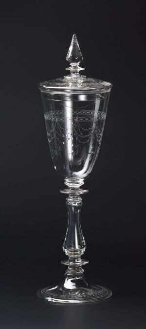  A tall, clear glass goblet with intricate etchings on a dark background, featuring a wide bowl, slender stem, and a pointed lid on top.