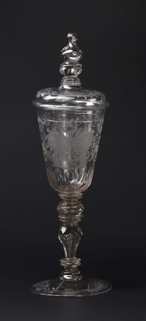  An intricately detailed clear glass goblet with an ornate lid finial and engraved patterns throughout, displayed against a dark background.