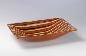  An elongated, boat-shaped ceramic bowl with layered bands of terracotta, off-white, and deep brown, displaying a polished surface that suggests careful craftsmanship. The piece exudes a sense of earthy elegance against a neutral light grey background.