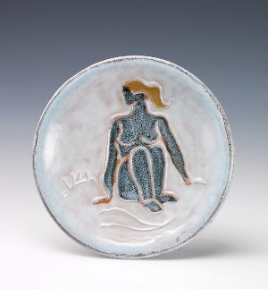  A handcrafted ceramic plate with a pale bluish-white glaze featuring a central raised relief of a stylized human figure in blue with ochre head and arms, and additional abstract design elements suggesting a natural setting.