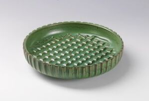  A circular, textured ceramic dish with a glossy green glaze, featuring a grid-like pattern in the center and vertical ridges along the edge. The dish is set against a plain background with a slight shadow indicating a light source to the side.