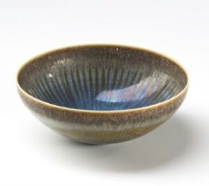  A ceramic bowl with a dark earthy rim leading into a lighter base on the exterior, and a vibrant, grooved interior with a gradient from mossy green to central azure blue, placed against a light background. Artist and title unknown.