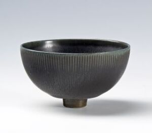  A lustrous deep charcoal-black ceramic bowl with fine, vertical ridges on its exterior, contrasting with a smooth interior and an unglazed, brown base on a light gray background.