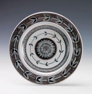  A ceramic plate with a white base, featuring intricate black sunburst and leaf-like radial patterns, displayed against a gray background. Artist name and title are unknown.