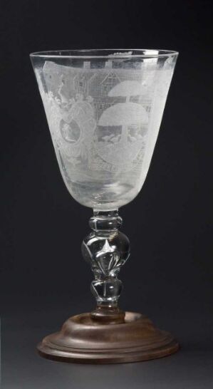  An elaborately etched glass goblet with a spiraled stem and a dark wooden base set against a dark background that emphasizes the craftsmanship and detail in the glass.