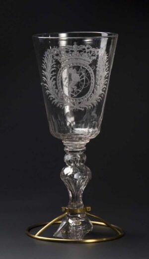  An intricately designed clear glass goblet with a monogram and leafy patterns etched on the bowl, a sculptured stem, and a metallic starburst base against a dark background.