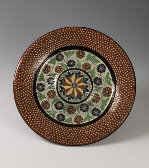  A decorative circular plate featuring an elaborate pattern with concentric circles. The outer rim is reddish-brown with a dotted motif, followed by a dark band, a ring of green with stylized red and blue flowers, and a central circle with a wheel-like design in blue, yellow, red, and turquoise.