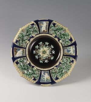  A decorative plate with a dark blue background and gold rim, featuring intricate green and white floral designs interspersed with gold patterns and a few red accents, centered by an ornate gold and green motif on blue.