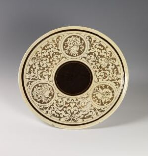  A decorative plate with dark brown and gold designs, featuring a central dark brown circle and an ornate gold pattern with floral motifs around it, set against a plain grey background. The artist's name and title are unknown.