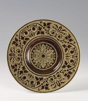  Decorative ceramic plate with intricate embossed floral and geometric patterns in caramel and dark brown colors, displayed against a neutral background. Artist name and title unknown.