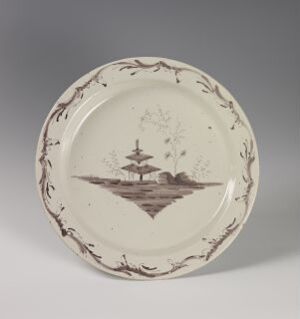  A slightly angled ceramic plate with a cream base color features a brown monochromatic landscape scene in the center, with a multi-tiered fountain or pagoda, trees, surrounded by a scalloped border adorned with vine-like twig patterns.