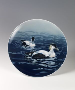  A circular plate featuring a painted image of two eiders floating on water with various shades of blue and white, illustrating a serene natural scene.
