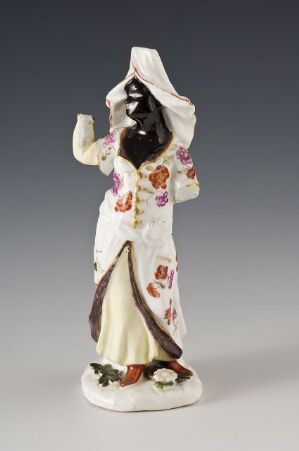  A porcelain figurine of a woman in a traditional dress with a floral pattern, standing gracefully with a raised hand and wearing a black headscarf that covers her face, set against a plain grey background.