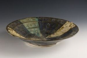 
 An ancient ceramic bowl with a broad, flat rim and a shallow basin, featuring a quadrant pattern with alternating turquoise and dark brown/black colors, displayed against a gradient background from light gray to black.