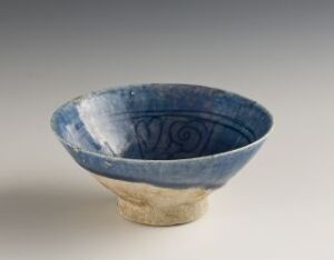  A handcrafted two-toned ceramic bowl with the upper section glazed in a vibrant cobalt blue featuring a swirling pattern and the lower section in natural beige clay, against a neutral background. Artist and title are unknown.