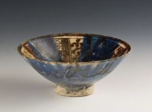  An antique bowl with a flared rim and footed base, featuring a mosaic of deep to light blue tones and gold accents within, set against a neutral gray backdrop. Artist name and title are unknown.