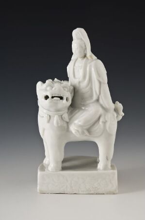 
 A porcelain sculpture of a seated woman in flowing robes beside a fluffy dog, both rendered in pure white against a neutral gray background.