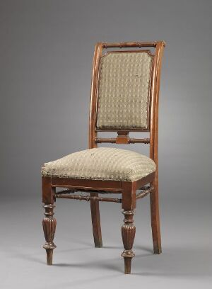  An antique wooden chair with a high back and upholstered seat in creamy beige fabric, featuring turned legs with decorative carvings, standing against a plain gray background.