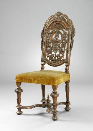  An ornate antique chair with a highly detailed, gilded backrest featuring symmetrical carvings and a velvety golden-yellow cushioned seat, set against a neutral background.