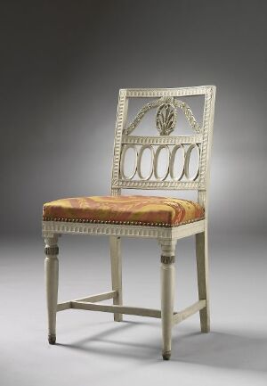  An elegant white-painted beechwood chair called "Nollstolen" featuring intricately carved designs on the high backrest with a rosette centerpiece and symmetrical oval spindles, and a plush seat covered in a golden-amber artificial silk fabric. The chair stands against a gradient gray background that highlights its design and craftsmanship.