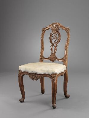  An antique-style wooden chair with elegantly curved legs, creamy white upholstered seat, and intricately carved openwork backrest set against a neutral gray background.