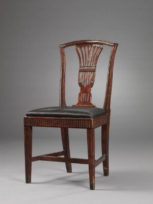  An antique-style wooden chair with a decorative vertical splat on its backrest is displayed against a subdued grey background. The chair has a dark, polished cherry wood finish and features intricate carvings along the seat's front edge.