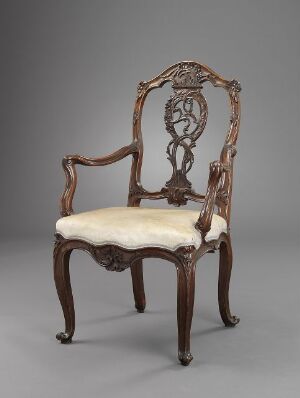  An antique wooden armchair with ornate openwork carving in the backrest, standing on elegant curved legs, and featuring a plush cream-colored seat cushion. The chair is against a grey backdrop, emphasizing its rich brown hues and elaborate design.