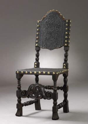  An antique-style high-backed chair with a detailed, embossed backrest and a frame of dark-toned wood, adorned with metallic studs, intricate carvings, and turned legs against a neutral gray background.