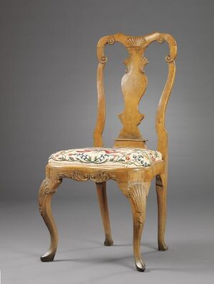  An antique beech wood chair titled "Stol" by an unidentified producer, with a high, intricately carved backrest and upholstered seat featuring colorful embroidered floral patterns on wool piqué fabric, against a neutral backdrop.