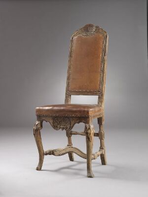  An antique, ornately carved wooden chair with a tall, curving backrest upholstered in faded light brown velvet, set against a simple grey background.