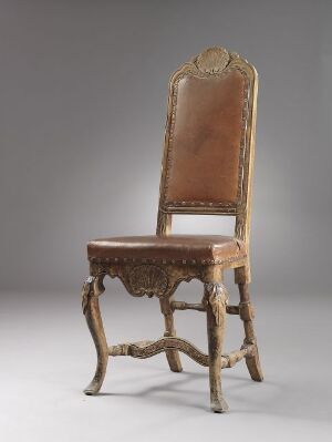  An antique wooden chair with a curved crest rail, a faded tan leather backrest and seat, and elaborate baroque-style carvings on the legs and apron, set against a graduated gray to white background.